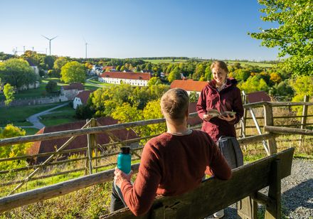 Persoon op een bankje, rechts staande persoon met flyer, op de achtergrond kloostercomplex