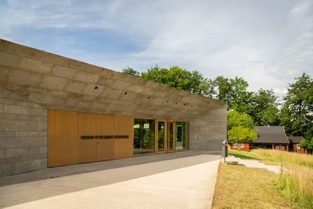 Modernes Museumsgebäude mit Betonfassade, großen Holzfenstern; im Hintergrund Bäume und ein Holzhaus.